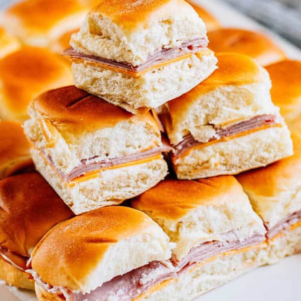 A close-up of several ham and cheese sliders on fluffy buns, stacked on a white platter.