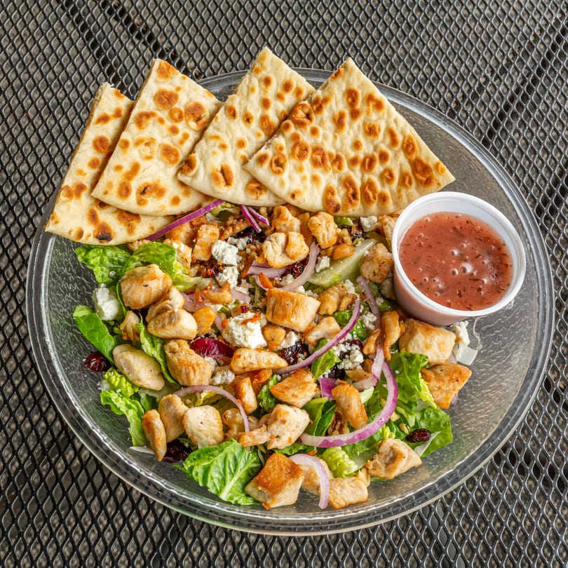 A clear bowl of chicken salad with lettuce, red onion, feta, and dried cranberries, served with pita bread and dressing.
