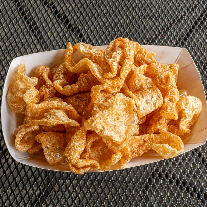 A white paper tray filled with golden-brown, crispy pork rinds on a dark metal mesh table.